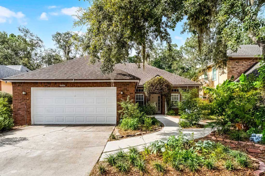 A single-story red brick home with a curved concrete walkway, framed by a mature live oak canopy with Spanish moss. The traditional front yard features a layered, ecologically diverse garden with lush green plantings, native shrubs, ornamental grasses, and dense foliage instead of a standard turf grass lawn.